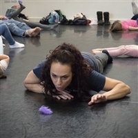 Woman with long dark curly hair wearing navy blue t-shirt lying on floor blowing a purple feather.